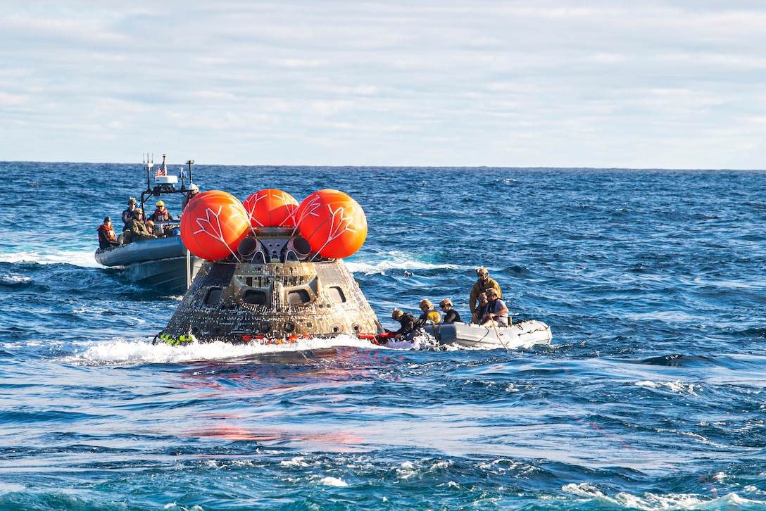 Sailors riding in small boats attach a line to a spacecraft in a body of water.