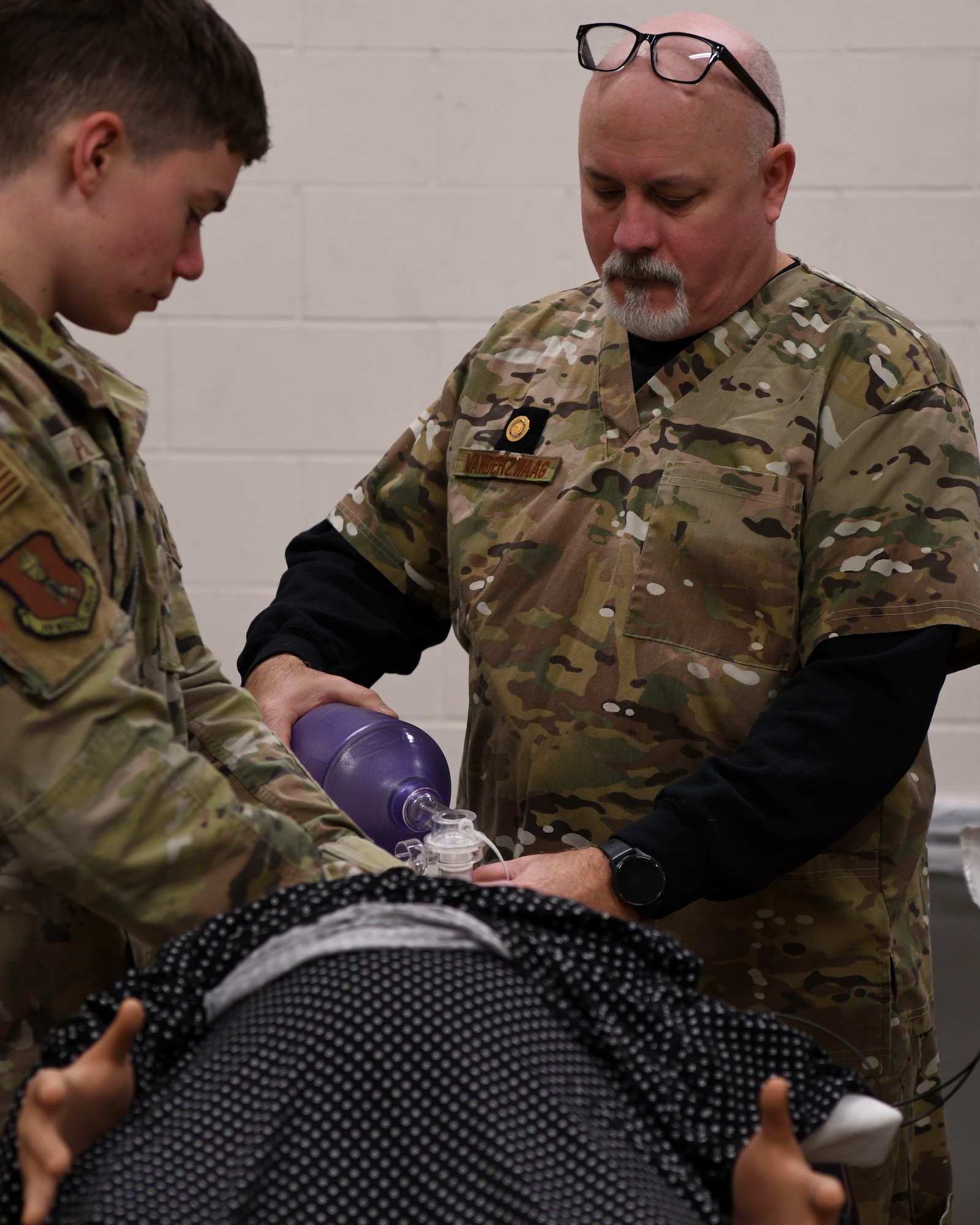 two men in green uniforms preform CPR on a mannequin.