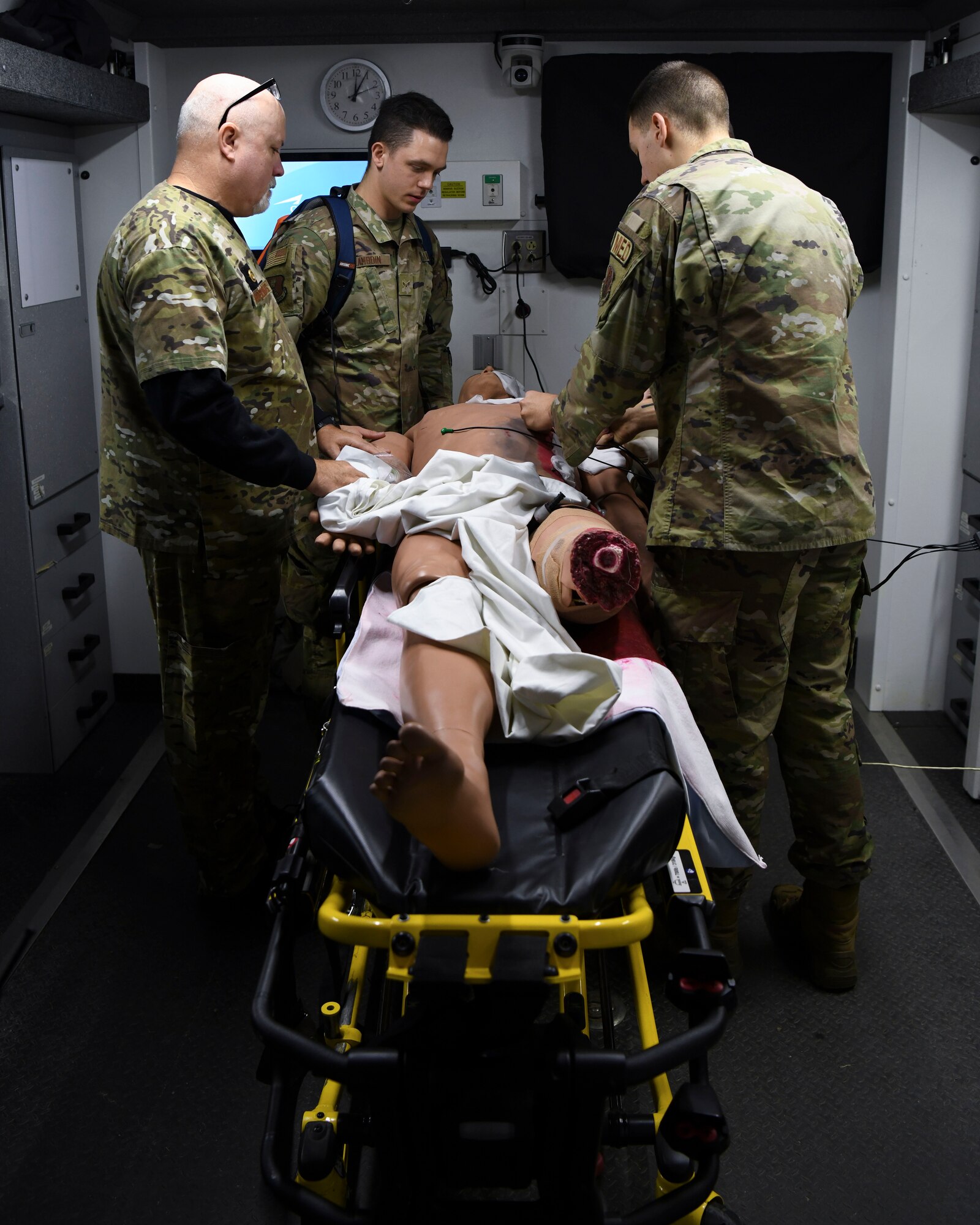 The men in green uniforms stand around a mannequin with injuries in the back of a simulated ambulance.