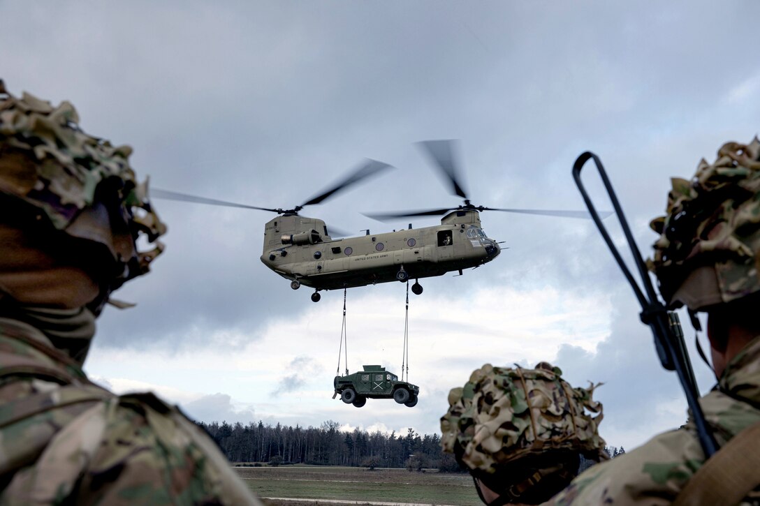 A military vehicle hangs by cable from a helicopter.