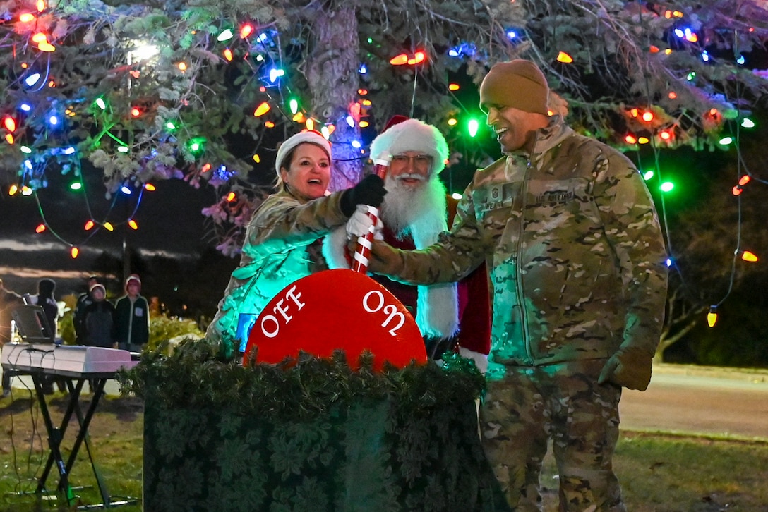 Two airmen and Santa push a giant red-and-white switch to "on" in front of a lit holiday tree outdoors.