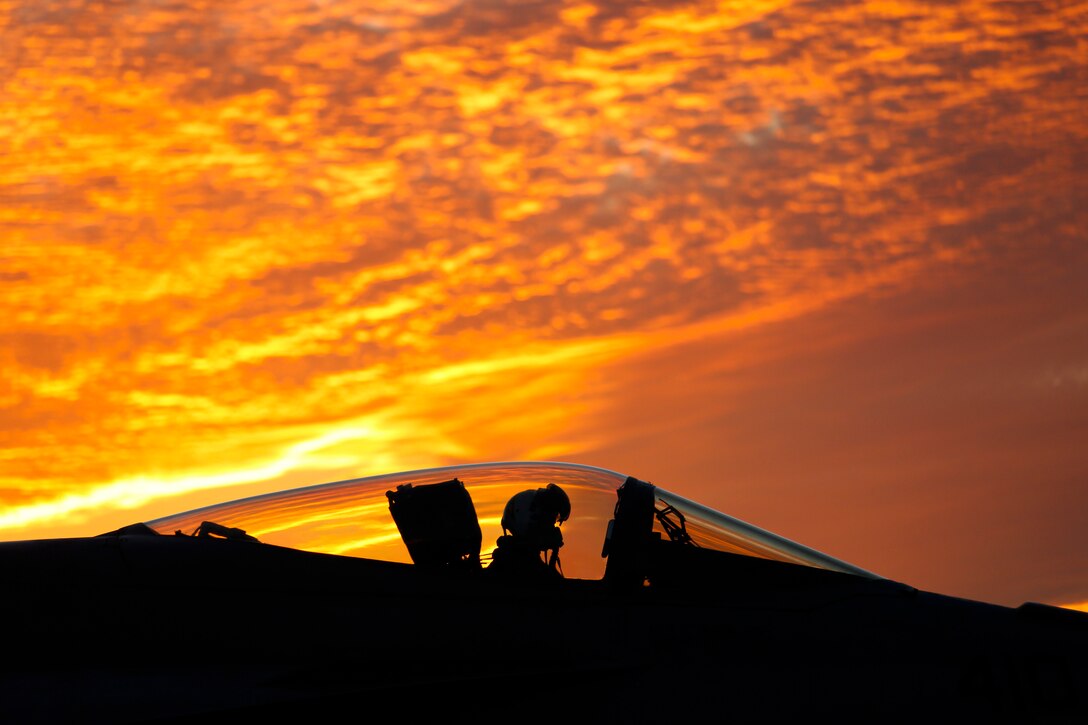 A pilot in an aircraft is shown in silhouette and profile against a brilliant orange sky.