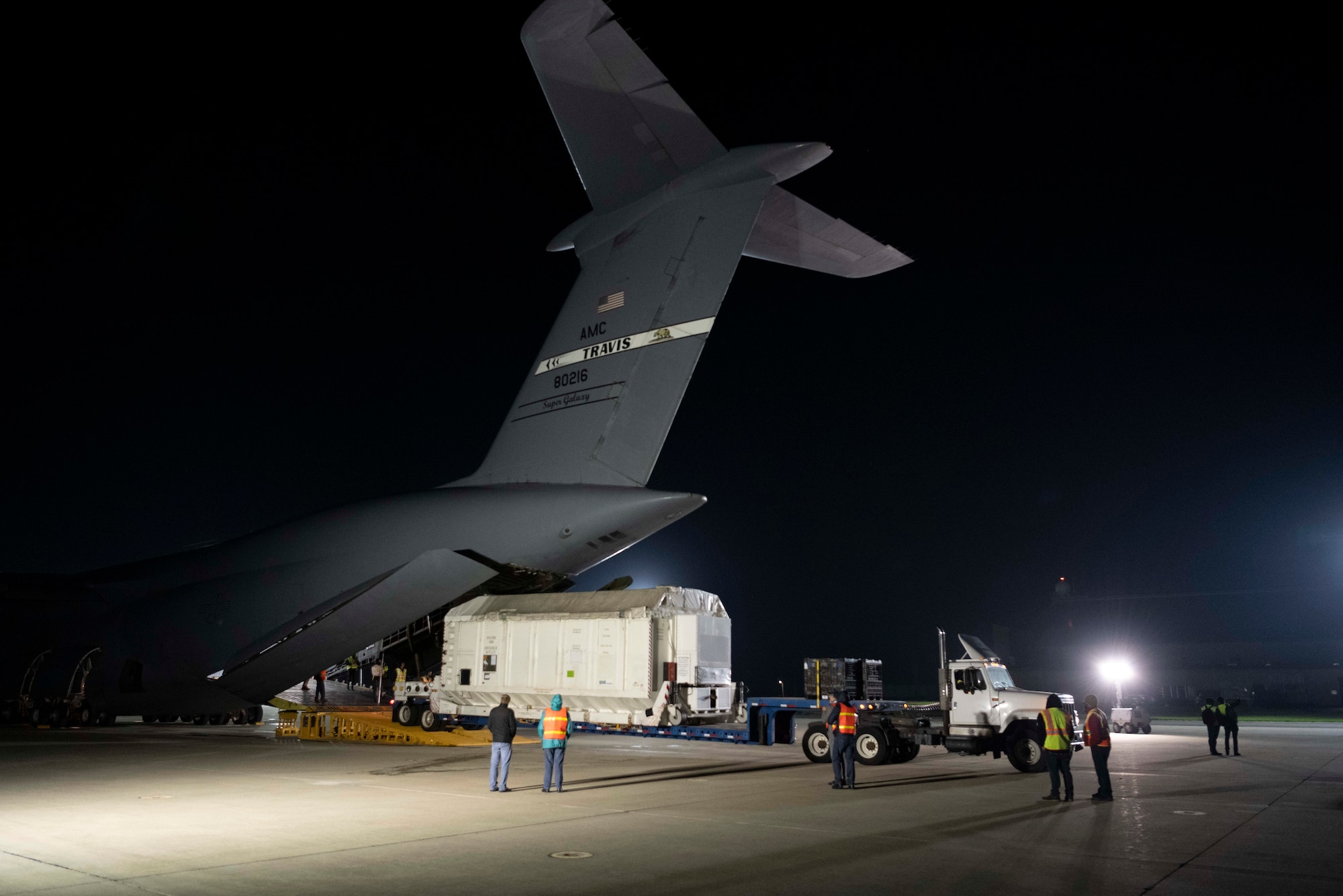 People move a satellite attached to a semi truck out of the cargo hold of a large aircraft
