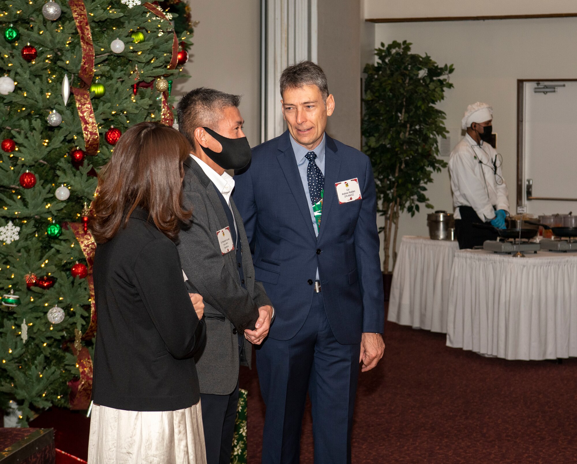 three people standing next to a Christmas tree talking with each other.