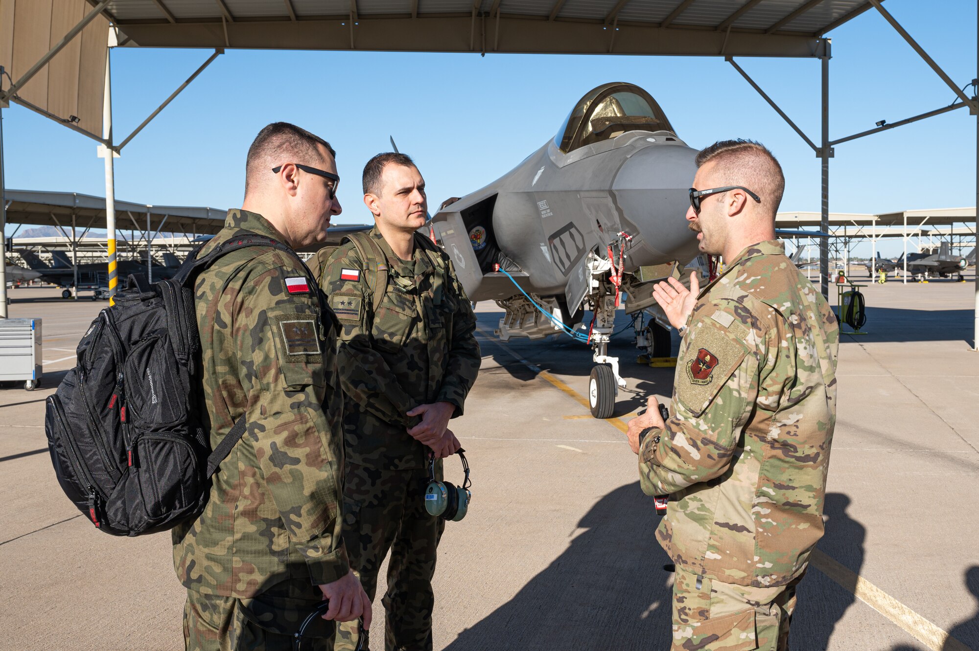 U.S. Air Force Tech. Sgt. Raymond Lemmert, 62nd Aircraft Maintenance Unit F-35 Lightning II crew chief (right), briefs Polish Air Force Lt. Col. Pawal Muzyczuk, logistics officer (left), and PAF Lt. Col. Rafal Zawadka, specialist officer (center), on F-35 maintenance operations Dec. 9, 2022, at Luke Air Force Base, Arizona.