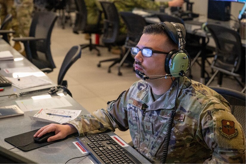 An airman looks forward while sitting at a long desk wearing headphones with a microphone.