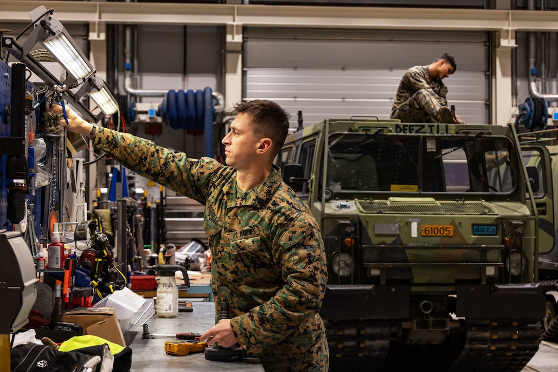 A Marine reaches for tools in a garage-type space while another works atop a vehicle in the background.