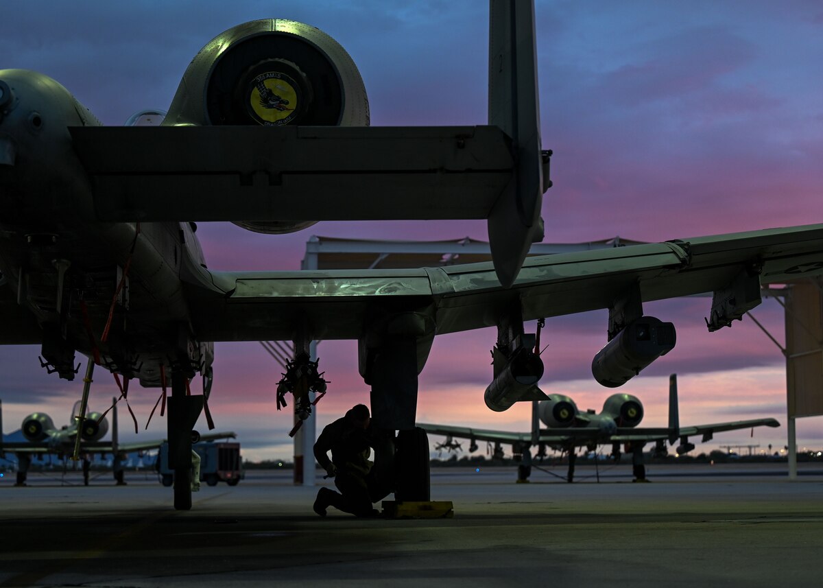 A photo of a man in a military uniform working on a military aircraft as the sun sets in the background.