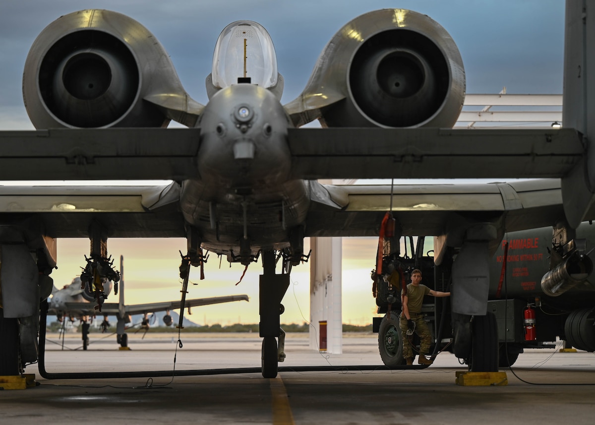A photo of a man in a military uniform refueling a military aircraft as the sun sets in the background.