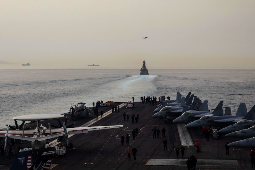 Sailors and aircraft are visible on a ship's deck as it travels in a body of water behind another ship.