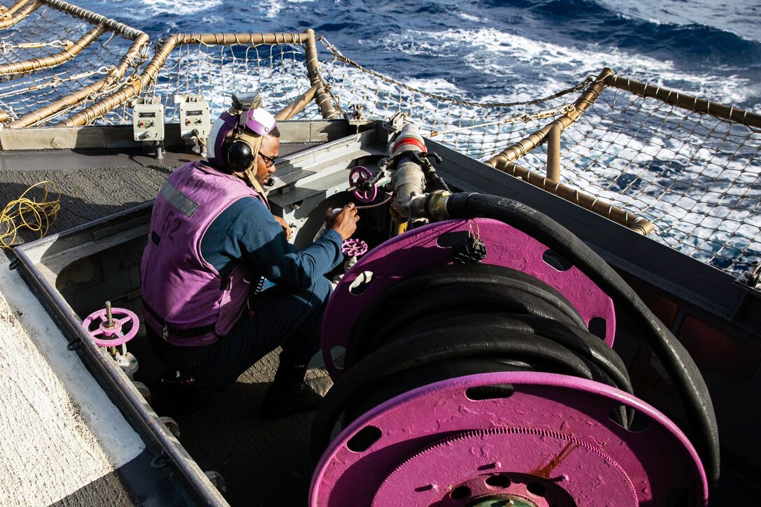 A sailor sits next to a fuel hose on a ship.