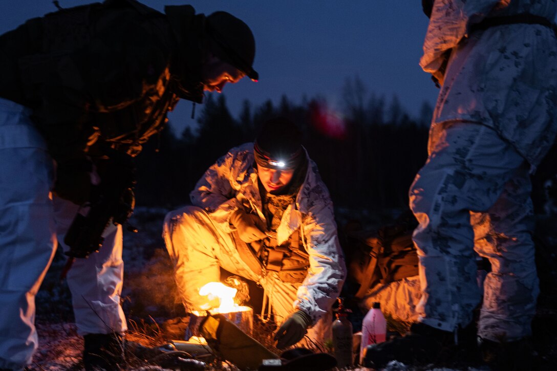 A Marine kneels while lighting a stove at night while two others look on.