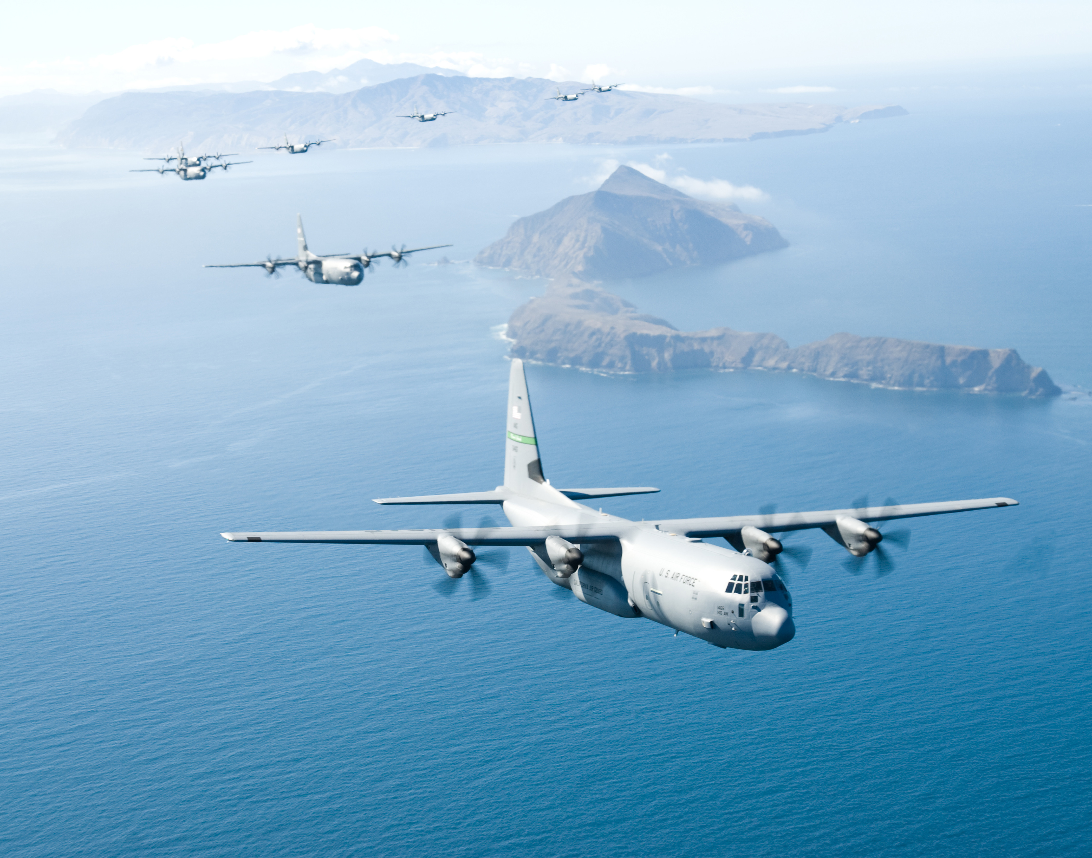 A photo of multiple military C-130J aircraft flying above an island in the ocean affixed with a large blue body of water.