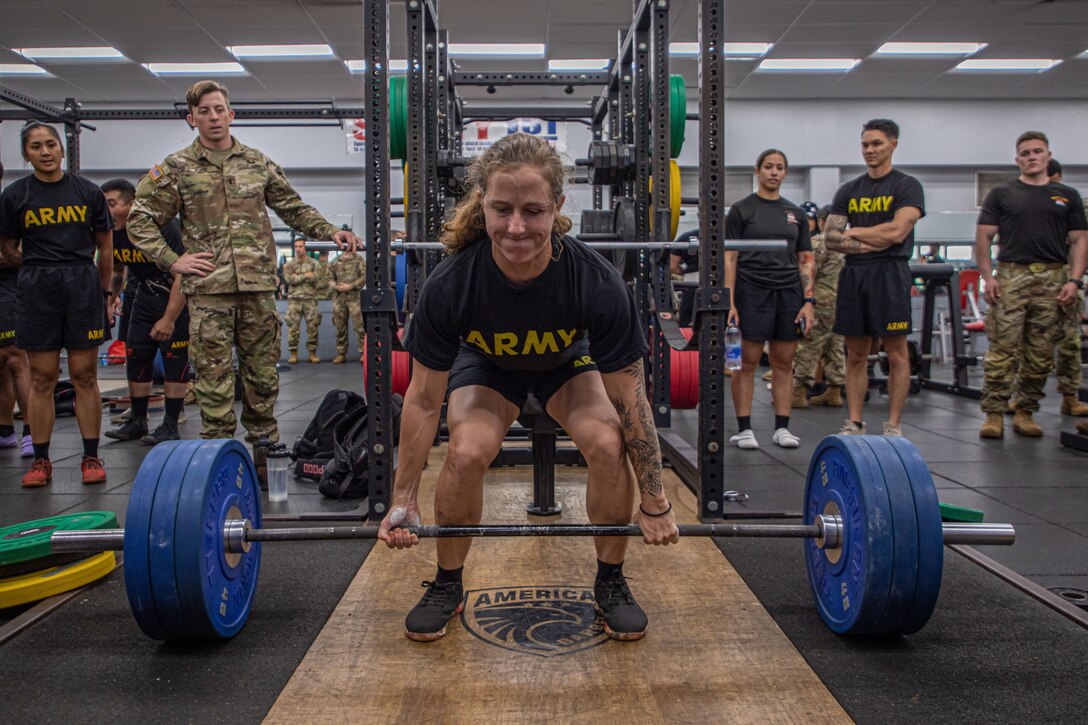 A soldier lifts a heavy barbell as others watch closely.