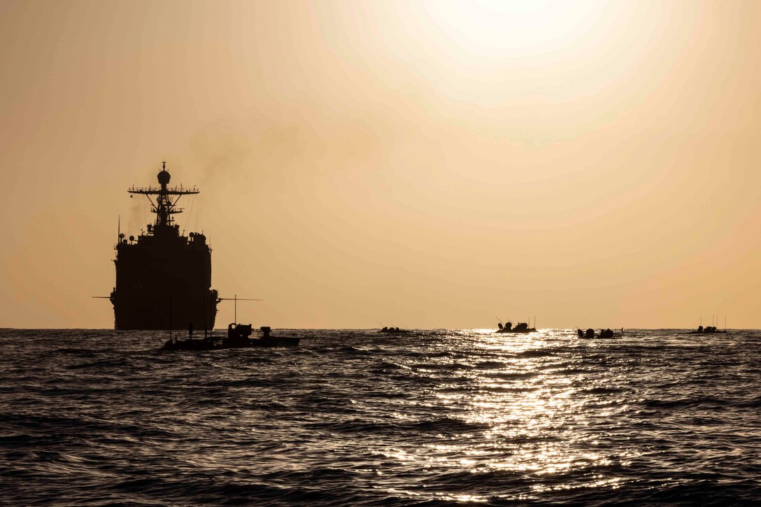 A ship and small boats shown in silhouette travel through waters.