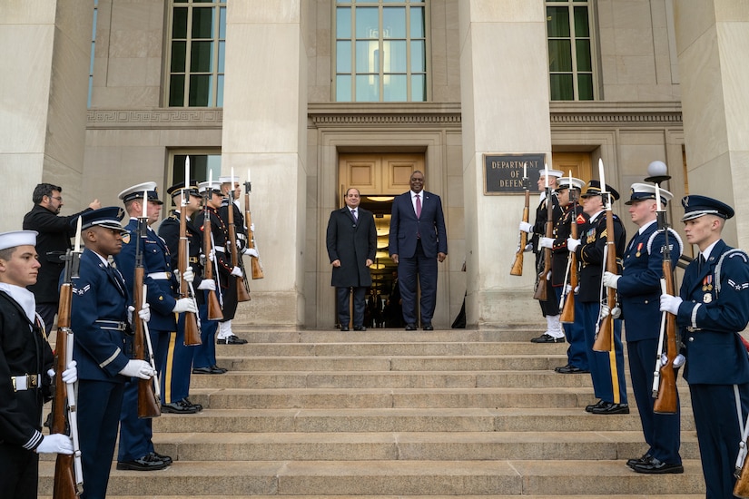 Two men stand at the top of stairs. Servicemembers in dress uniform are lined up on either side along the stairs.