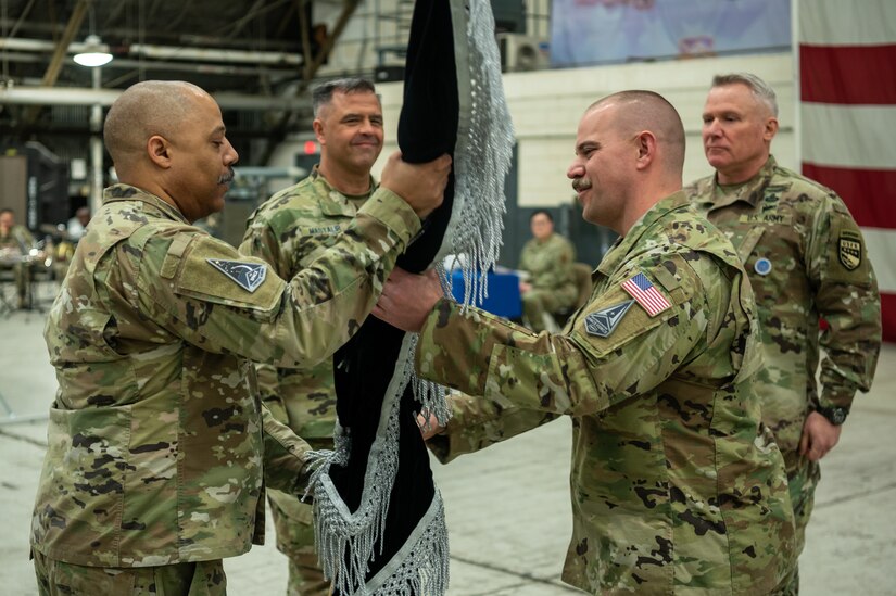 Two service members pass a guidon from one to another while two other service members stand at attention.