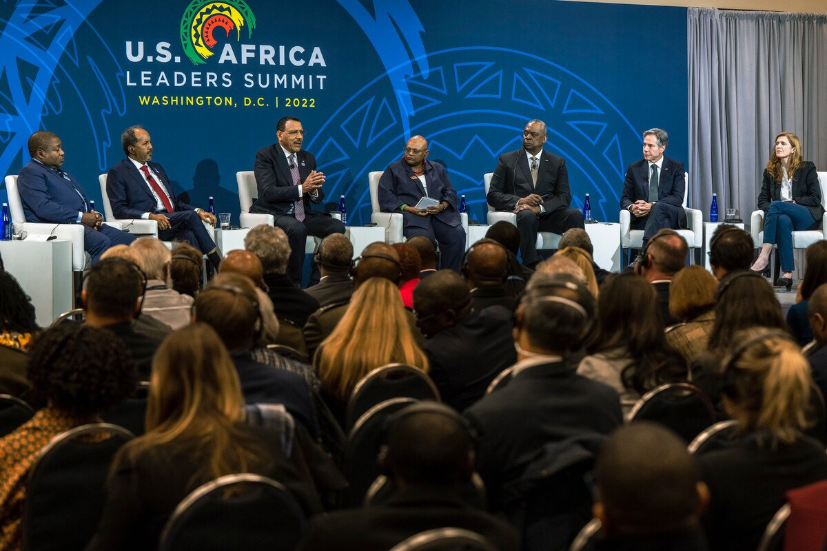 In a crowded room, seven individuals are seated on a stage.
