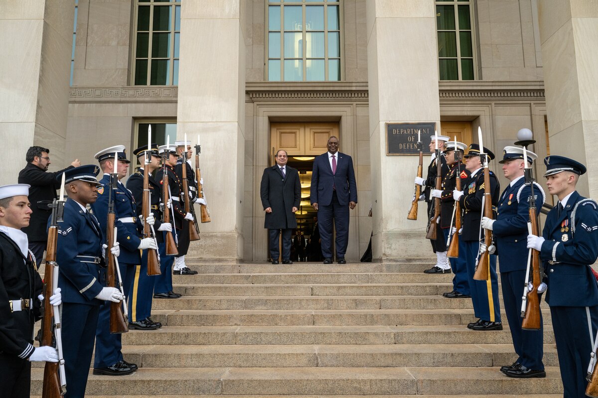 Two men stand at the top of stairs. Servicemembers in dress uniform are lined up on either side along the stairs.