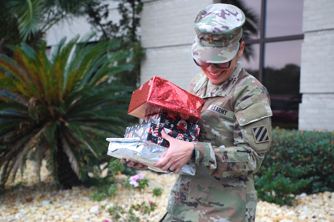 A smiling soldier carries a pile of wrapped gifts in her arms.