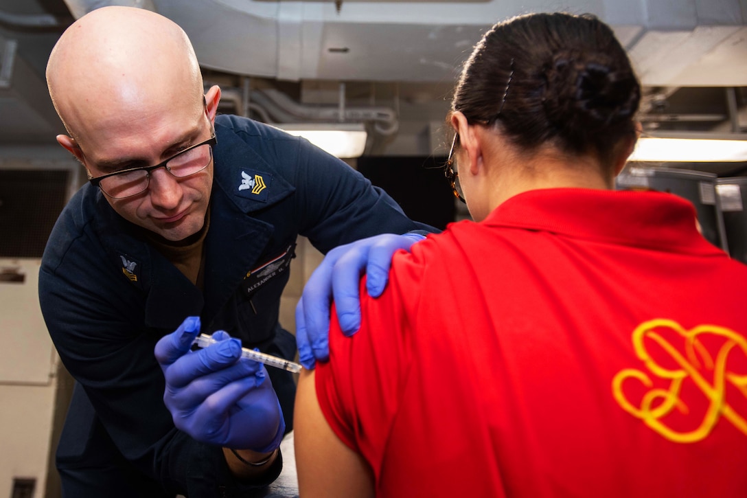 A sailor wearing gloves holds a syringe while administering a COVID-19 vaccine booster shot.