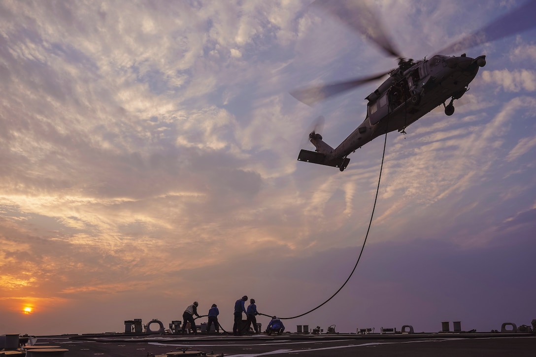 Sailors aboard a ship at sea hold a fuel line attached to a hovering helicopter under a sunlit sky.