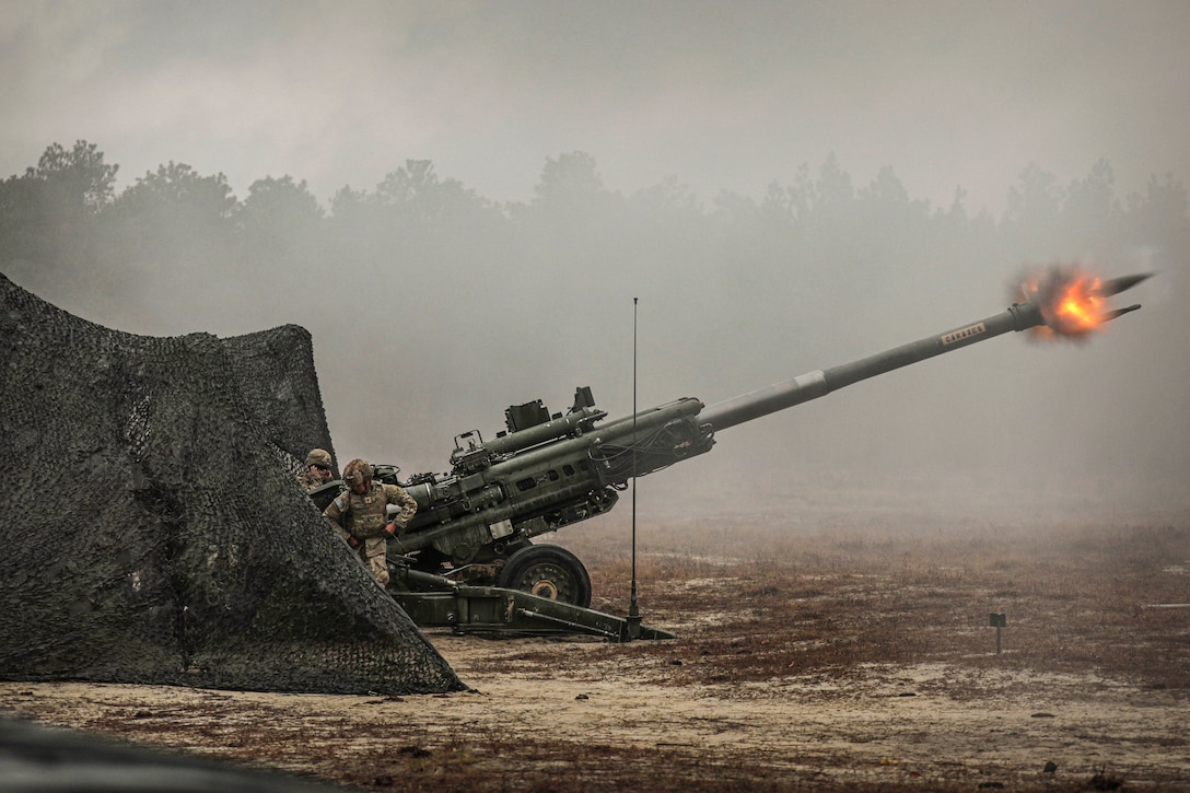 Soldiers fire a weapon in a field near a tent.