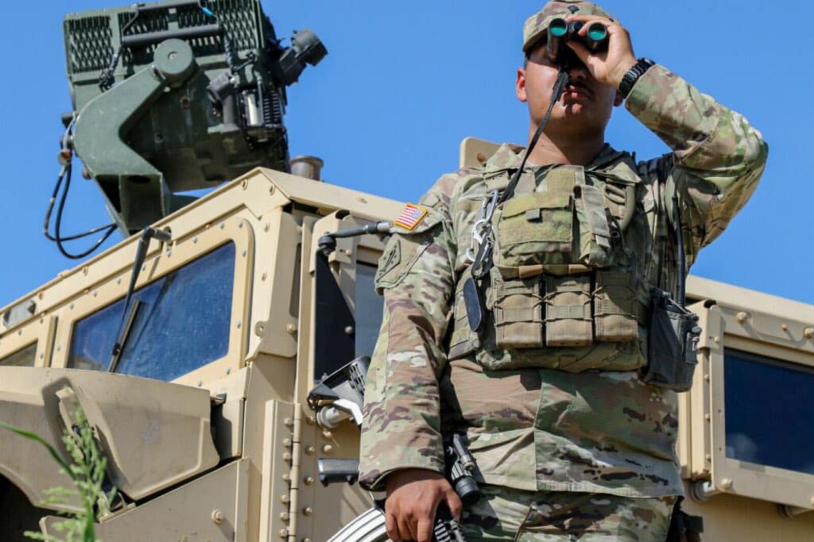 Army Guardsman looks through binoculars standing by a humvee.