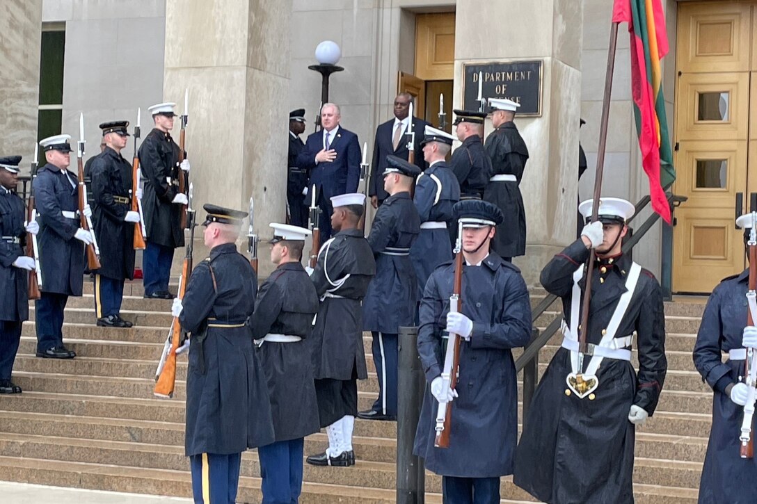 Two men stand outdoors at the top of stairs; military members stand along the stairs at attention.