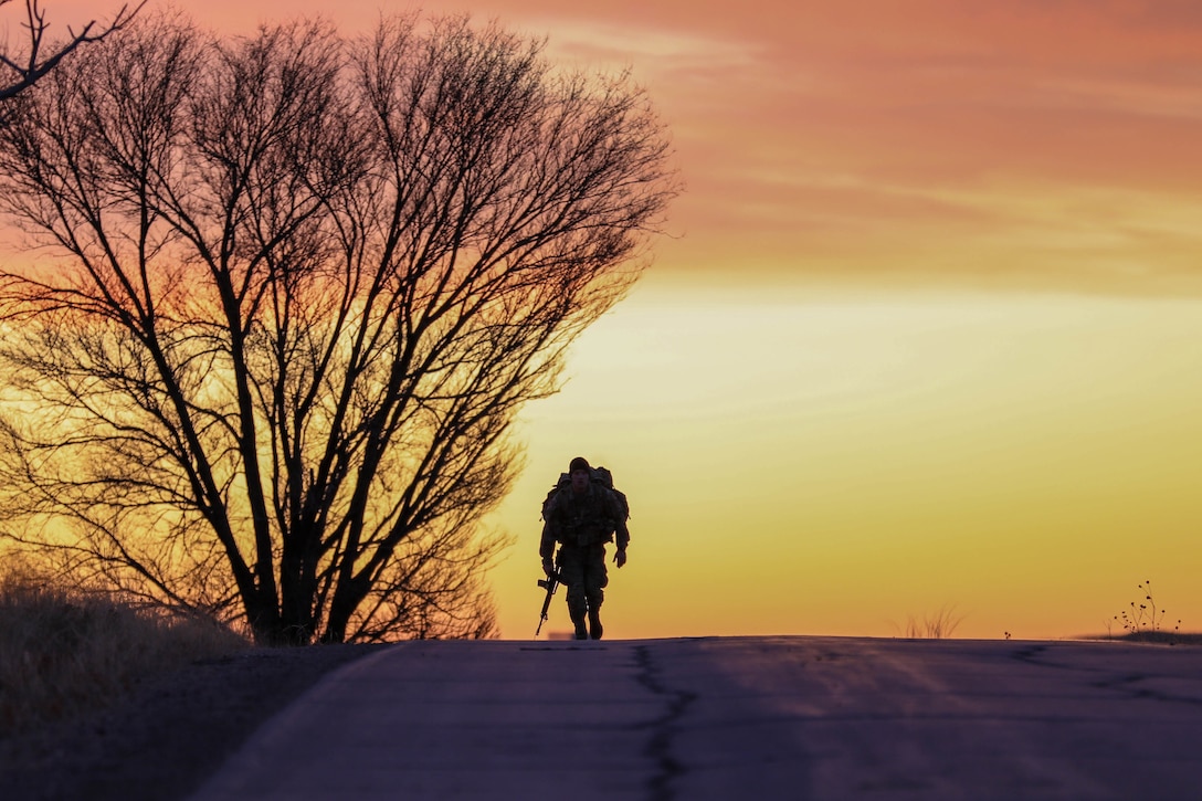 A soldier walks along a road at twilight carrying a weapon and backpack.