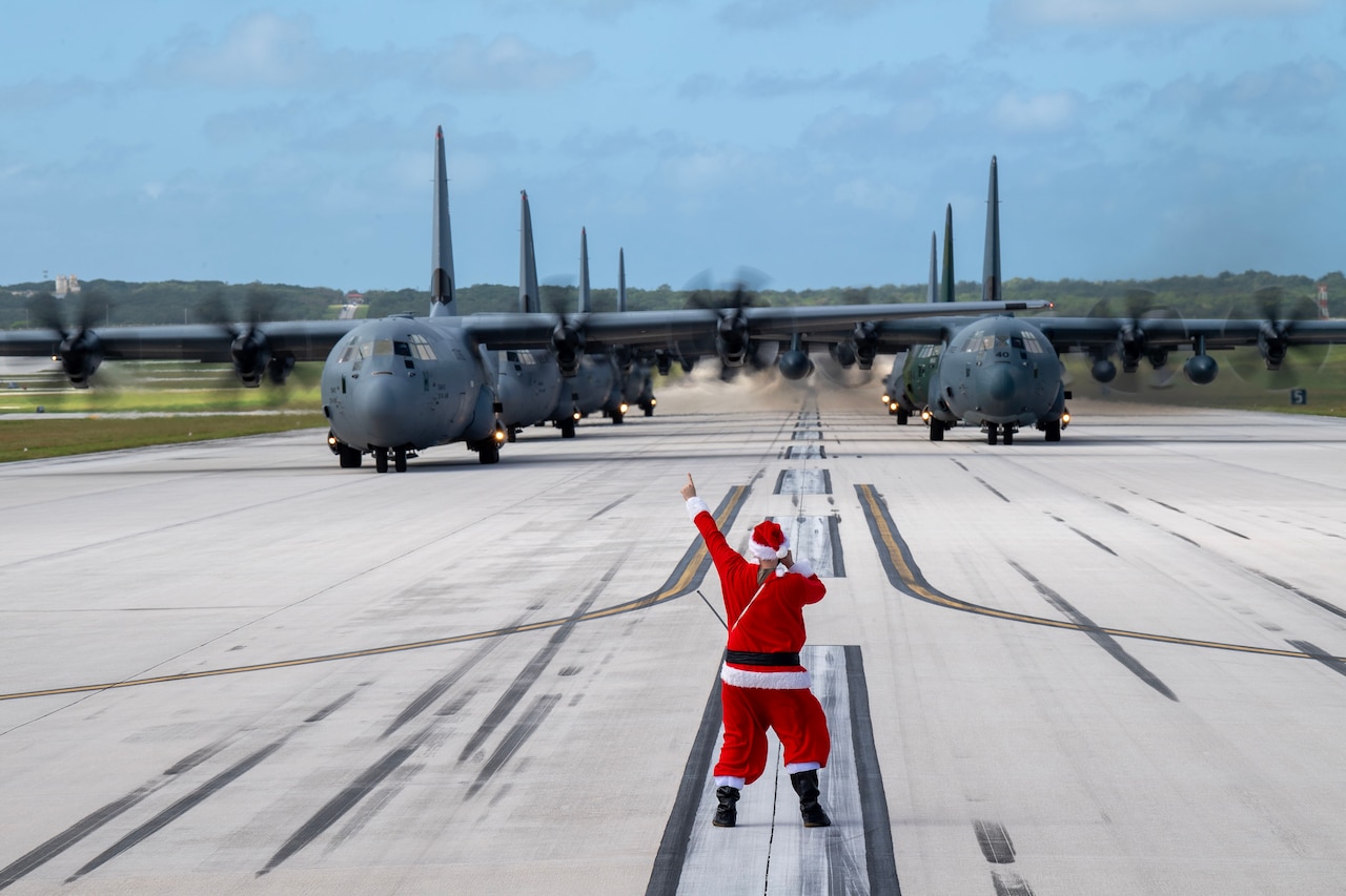 An airman dressed as Santa Claus guides rows of large military aircraft on a runway.