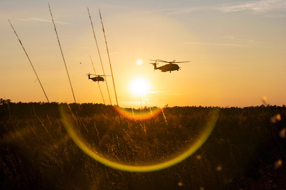 Two helicopters fly above a field under a sunlit sky.