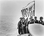 Coast Guardsmen pose in front of a USCG ensign and commissioning pennant.  They are equipped as a landing force.