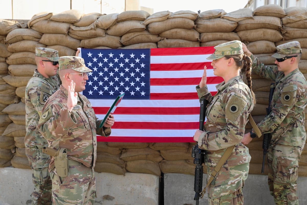 Two soldiers face each other raising their right hands in front of an American flag.