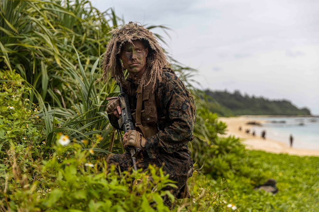 A Marine dressed in camouflage walks through a jungle terrain near a beach.