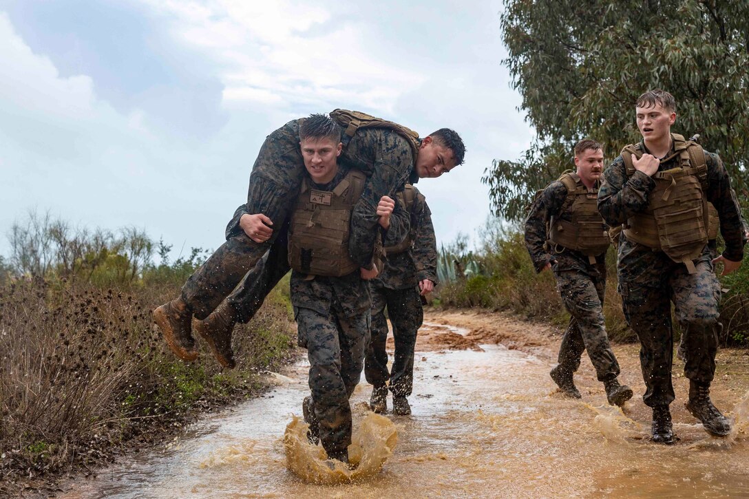 A Marine carries another Marine while walking through a flooded trail; other Marines walk beside them.