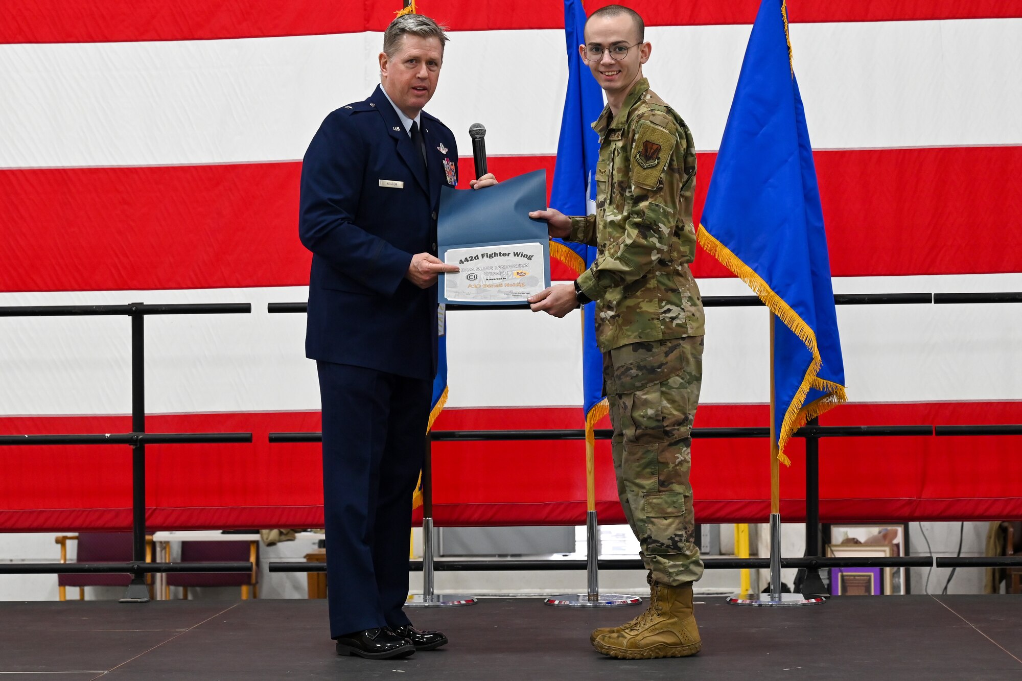 A man in a blue dress uniform hands a certificate to a man in camouflage fatigues.