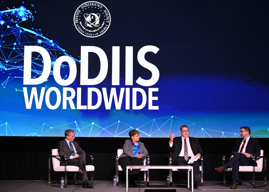 Image of four people sitting on a stage with text behind that reads. DoDIIS Worldwide, in white letters on a blue and black background with the DIA Logo above.