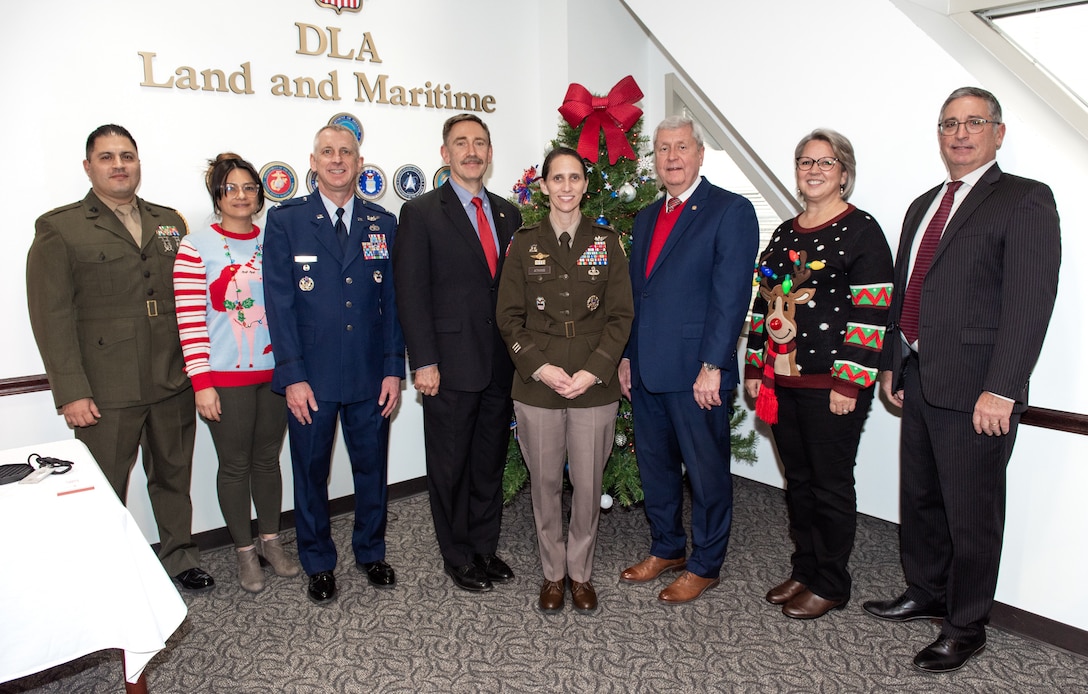 Five men and three women pose for a photo in the corner of a room. One man is in a olive green Marine Corps dress uniform, another man is in a Air Force dress blues uniform and the dark haired woman in the center wears a olive green Army dress uniform. The other two women wear Christmas sweaters and the other three men are wearing formal suits with ties. Two are black and one is dark blue.