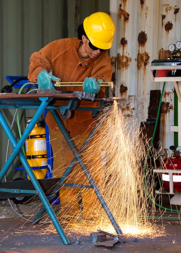 A person in protective equipment uses a cutting torch on metal. the torch is creating lots of orange sparks that are falling toward the deck and away from the person cutting.