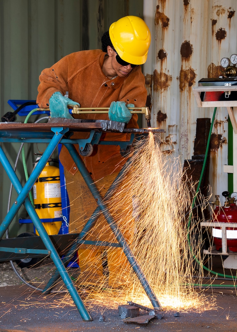 A person in protective equipment uses a cutting torch on metal. the torch is creating lots of orange sparks that are falling toward the deck and away from the person cutting.
