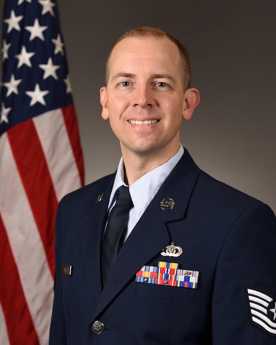 An official head shot photo of TSgt Travis Hyde in front of the American flag. He is wearing the blue service dress uniform.