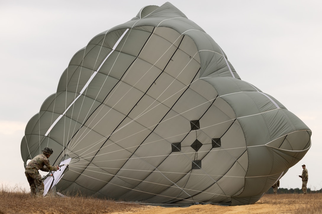 A soldier pulls in an open parachute by the strings while another observes from the other side in a field.
