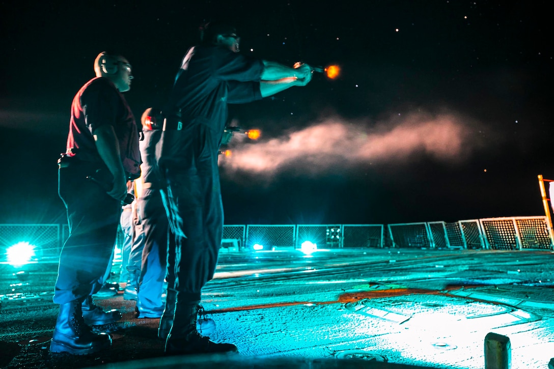 Sailors fire weapons aboard a ship at sea in the dark as a fellow sailor watches.