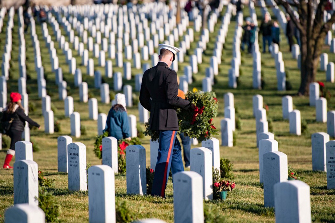 A service member and volunteers walk through a cemetery carrying wreaths.