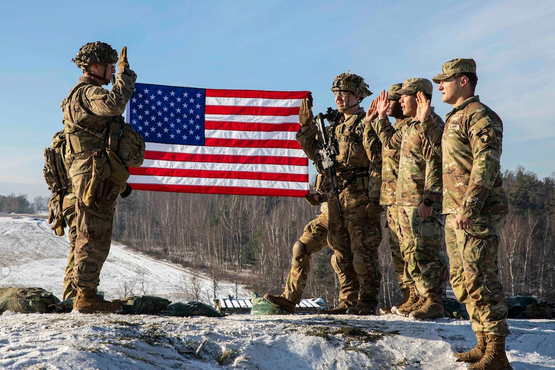 A soldier holds up his right hand across from other soldiers holding up their right hands in front of an American flag.