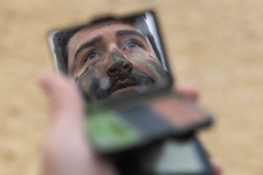 A Marine covers his face with camouflage paint as seen through a small mirror.