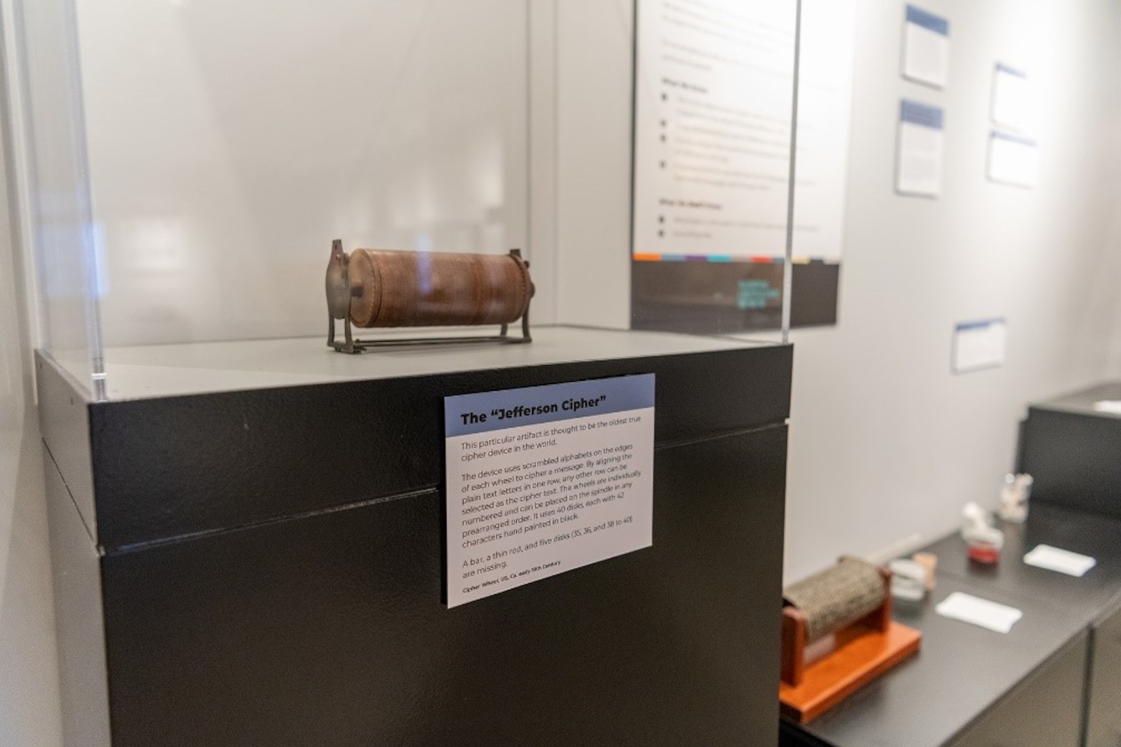 An image of the “Jefferson Cipher," a small brown cylinder mounted on four legs, displayed in a glass case