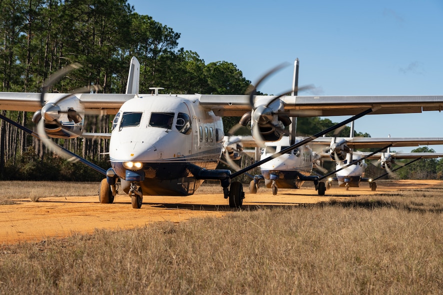 Three smaller aircraft with spinning propellers are parked in a straight line along a small dirt taxiway with grass in the foreground and trees in the background. A slight orange tint is reflected by the bottom of the airfoil from the dirt.