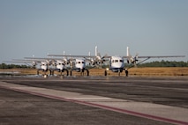 Four aircraft roll across cement in a straight line in close proximity to one another with a grassy backdrop